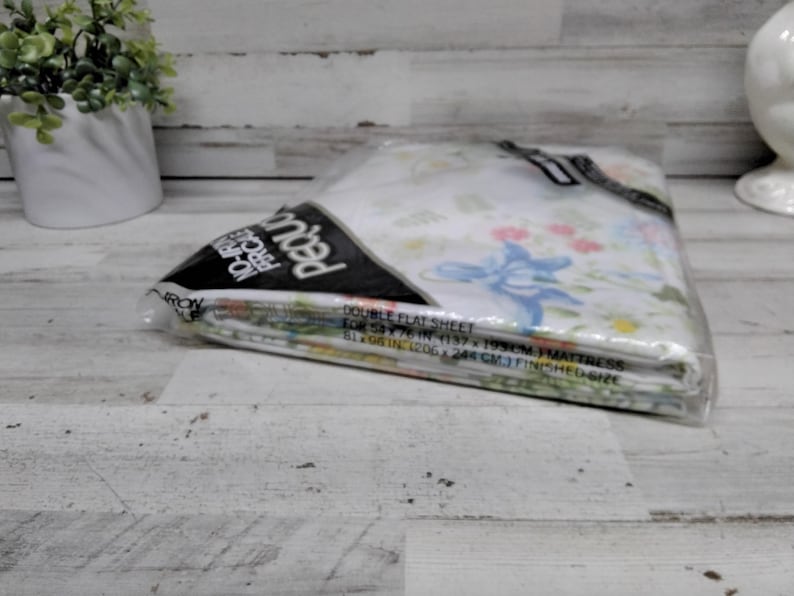 K&ouml;nnte beinhalten: Ein verpacktes Doppel-Flachbettlaken-Set f&uuml;r eine 137 x 193 cm Matratze. Das Bettw&auml;sche-Set befindet sich in einer transparenten Kunststoffverpackung. Die Bettlaken haben ein Blumenmuster mit blauen und rosa Blumen. Der Text auf der Verpackung lautet "No-Iron Percale".