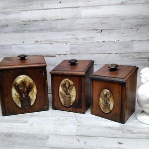 Pode incluir: Três potes de cozinha de madeira com tampas marrons escuras e incrustações decorativas ovais com desenhos de utensílios. Os potes são de tamanhos diferentes e têm botões marrons escuros. Um galo de cerâmica branca está à direita.