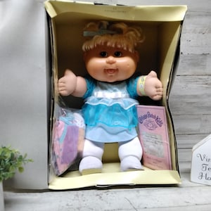 Puede incluir: Una muñeca Cabbage Patch Kids con cabello rubio y un lazo azul, vestida con un vestido azul claro con una cinta blanca. La muñeca está en una caja de cartón con accesorios envueltos en plástico y un folleto rosa. Un letrero dice Vintage Tenn House.