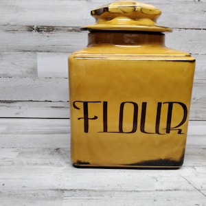 Puede incluir: Un tarro de cerámica amarillo para harina con tapa y la palabra "FLOUR" escrita en letras negras.