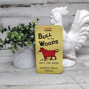 Puede incluir: Una lata de tabaco sin humo vintage "Bull of the Woods" con una ilustración de toro rojo sobre fondo amarillo. El texto dice "HE'S THE BOSS" y "Smokeless Tobacco Pleasure". Un gallo de cerámica blanco y una pequeña planta en el fondo.