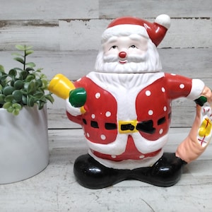 May include: A ceramic Santa Claus figurine, featuring a red suit with white polka dots and trim, a black belt with a gold buckle, and black boots. The figurine holds a yellow horn and a sack. The Santa Claus figurine is a festive holiday decoration.