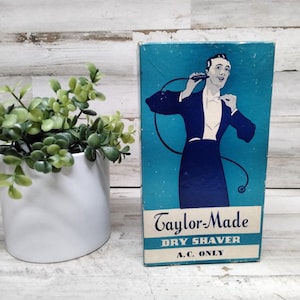 May include: Vintage Taylor-Made Dry Shaver box with a teal background and a man in a tuxedo. The box has the text "Taylor-Made Dry Shaver A.C. ONLY". A white ceramic pot with green plants is to the left.