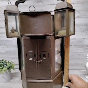Puede incluir: Una linterna antigua de metal marrón con asa y dos faroles laterales. El cuerpo principal tiene una puerta con recortes decorativos. La linterna mide aproximadamente 51 cm de alto.