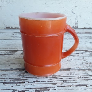 Puede incluir: Una taza de vidrio naranja con asa. La taza tiene forma cilíndrica con estrías horizontales y un interior naranja más claro. La taza está sobre un fondo de madera blanca desgastada.