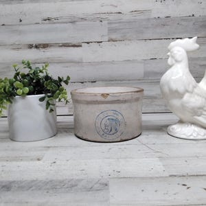 Peut inclure: Un coq en céramique blanche, un petit pot blanc avec du feuillage vert et un pot en grès vintage avec un emblème bleu sont disposés sur une surface en bois clair. Le coq est à droite, le pot à gauche et le pot au centre.