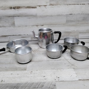 May include: A set of miniature aluminum kitchenware, including a teapot, a frying pan, and four cups. The set is made of shiny silver aluminum with black plastic handles.