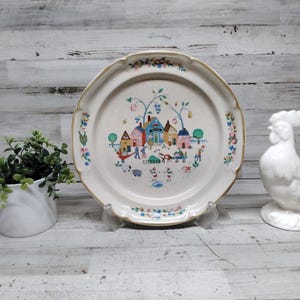 Puede incluir: Un plato decorativo color crema con una escena de pueblo en el centro. El plato tiene un borde dorado y detalles florales. También se ve un gallo de cerámica blanca y una pequeña planta en maceta.