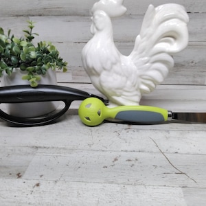 May include: A black kitchen tool with a handle and a curved design, a green and gray kitchen tool with a blade, and a white ceramic rooster figurine. A small potted plant is also in the image. The items are on a white wooden surface.
