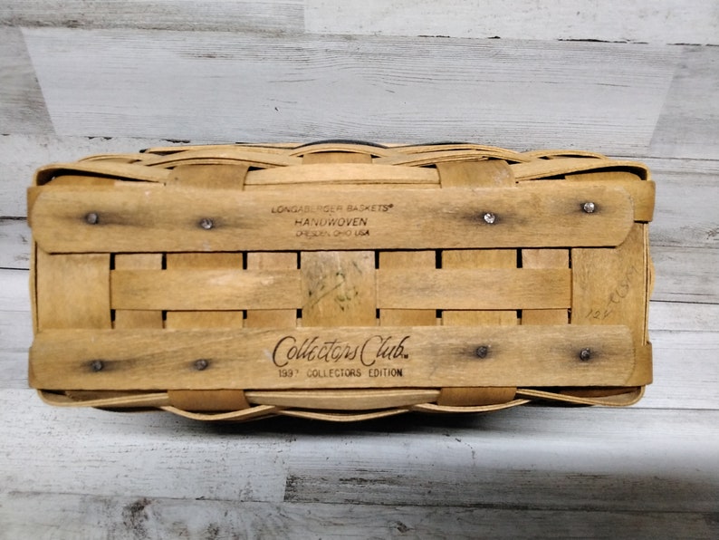 May include: Handwoven rectangular basket with a light brown finish. The bottom of the basket features the text "Longaberger Baskets Handwoven Dresden, Ohio USA" and "Collectors Club 1997 Collectors Edition". The basket is made of woven wood strips.
