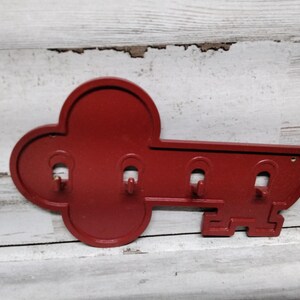 May include: A red key-shaped wall hook with three hooks for hanging items. The key is made of metal and has a distressed finish. The key measures approximately 10 inches long.