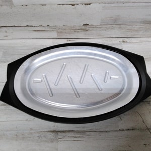 Puede incluir: Un plato para hornear ovalado de color negro y plateado con un borde elevado y una superficie texturizada. El plato tiene una serie de líneas paralelas grabadas en el fondo.