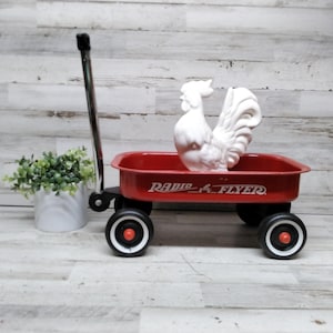 Könnte beinhalten: Ein roter Radio Flyer-Wagen mit schwarzen Rädern und einem silbernen Griff. Ein weißer Keramik-Hahn sitzt im Wagen. Links vom Wagen steht ein kleiner weißer Topf mit grünen Pflanzen.