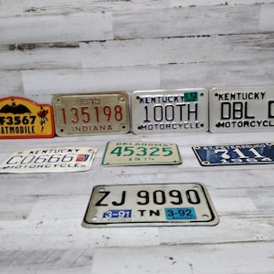 May include: A collection of vintage license plates, including Indiana, Kentucky, and Oklahoma plates. One plate is a Batmobile plate. The plates are in various colors and sizes, with visible numbers and text.