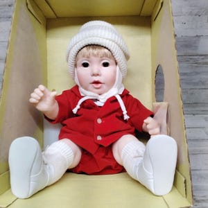 Op de afbeelding: Een vintage pop gekleed in een rode outfit, witte sokken en witte schoenen, zittend in een kartonnen doos. De pop heeft een witte gebreide muts en een lieve uitdrukking. De outfit van de pop bestaat uit een rode jas en bijpassende shorts.