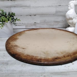 May include: A round wooden pizza stone or serving board with a light-coloured centre and a dark, weathered, distressed brown edge. The surface shows signs of use with visible scratches and wear marks across the circular wooden material.