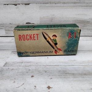 May include: A green and white box with the text "ROCKET A-1 TINY GERMANIUM RADIO" and a cartoon of a person riding a rocket.