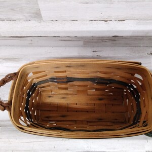 May include: A rectangular woven basket with a light brown wooden frame and a dark grey interior trim. The basket has two braided brown leather handles. The basket is empty and sits on a white wooden surface.
