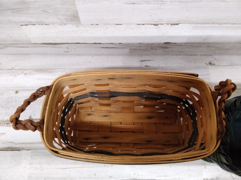 May include: A rectangular woven basket with a light brown wooden frame and a dark grey interior trim. The basket has two braided brown leather handles. The basket is empty and sits on a white wooden surface.