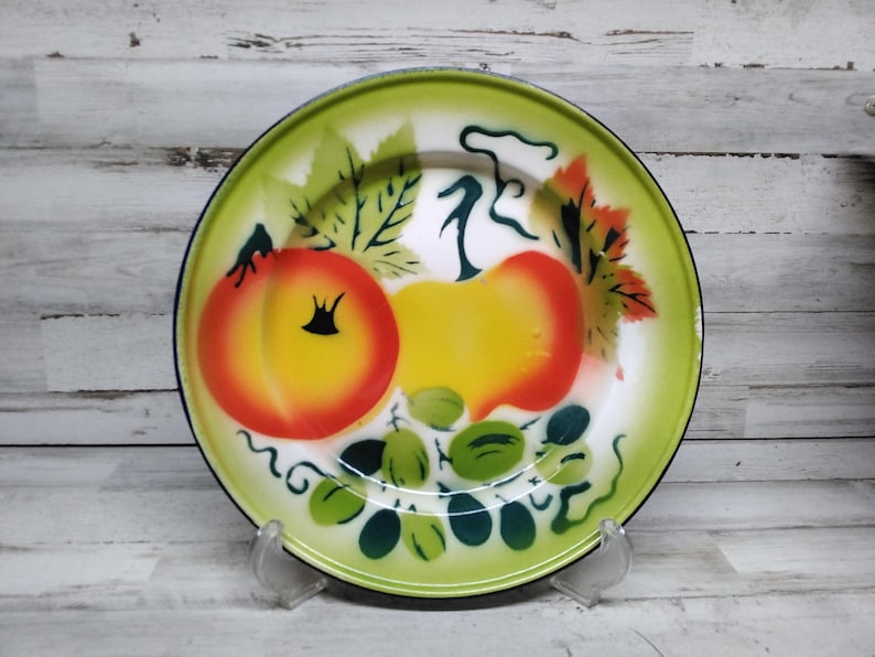 Peut inclure: Une assiette en &eacute;mail vert avec un centre blanc et un motif peint de fruits. Les fruits comprennent deux p&ecirc;ches jaunes et orange et des raisins verts. L'assiette a un bord noir.