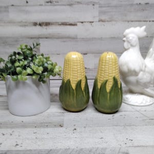 May include: A still life arrangement featuring a white ceramic rooster figurine, two corn-shaped salt and pepper shakers, and a small white pot with green foliage. The salt and pepper shakers are yellow with green husks.