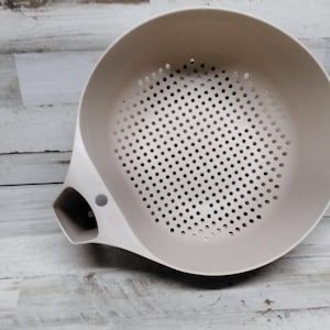 May include: A beige plastic colander with a handle and a small spout. The colander has a round shape and a mesh bottom.