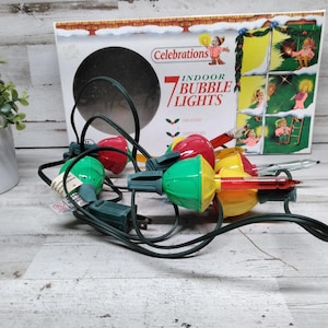 Op de afbeelding: Een set vintage Celebrations 7 Indoor Bubble Lights in een doos. De lampen zijn rood, groen en geel met een groen snoer. De doos heeft een feestelijk ontwerp met illustraties van mensen.