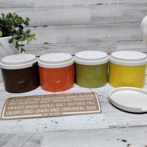 May include: Four colorful spice shakers with white lids, in brown, orange, green, and yellow. A white oval lid sits next to them. A label lists various spices. A white ceramic hen is in the background.