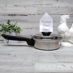 May include: A stainless steel saucepan with a black handle and a lid featuring a white tag printed with the text Vintage Tenn House. The pan is positioned next to a white ceramic rooster figurine and a small potted green plant.