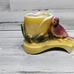 Puede incluir: Un macetero de cerámica amarillo con un diseño de pájaro. El macetero tiene una abertura en forma de tulipán y una base con forma de pata de pájaro. El pájaro es rosa y tiene un pico amarillo.