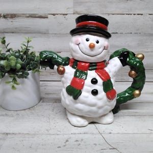 May include: A ceramic snowman teapot with a black top hat, red and green scarf, and a green wreath handle. The snowman has a smiling face with a carrot nose and black button eyes. A white ceramic pot with green plants is to the left.