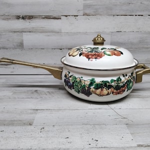 Puede incluir: Olla de esmalte blanco con asa y tapa de latón. La olla está decorada con un diseño colorido de frutas y flores.