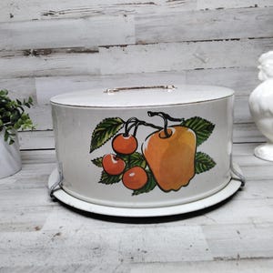 May include: A vintage white cake carrier with a lid and handle. The side of the carrier features a painted design of an orange apple and red cherries with green leaves. The carrier sits on a white plate and has metal supports.