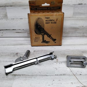 Peut inclure: Un accessoire de meulage de foret en argent avec une base en métal et une roue rotative. L'emballage indique "Adjustable Drill Sharpening Attachment" et "Fast, Accurate, Easy to Use".