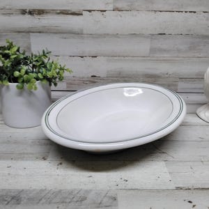 Puede incluir: Bol ovalado blanco con un fino borde verde. Una figura de gallo de cerámica blanca y una pequeña planta en maceta también están en la imagen. Los artículos están sobre una superficie de madera clara.