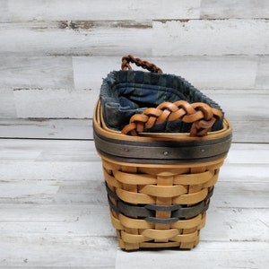 May include: A woven basket with a light brown body, dark grey bands and a braided leather handle. The interior has a blue and green plaid fabric lining. The basket has a square base and a rounded top.