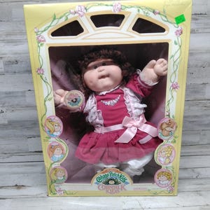 May include: A Cabbage Patch Kids doll in its original box. The doll has brown curly hair and is wearing a pink dress with white lace trim and a matching bow. The box is yellow with floral accents and the Cabbage Patch Kids logo.