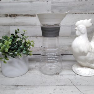 May include: A clear glass measuring container with a white funnel and gray accents. A white ceramic rooster and a small white pot with green plants are also in the image. The container has measurement markings.