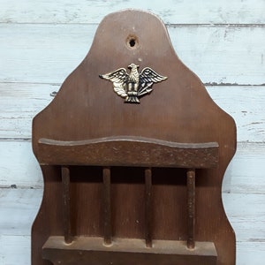 May include: Vintage wooden wall-mounted organizer with a decorative brass eagle emblem. Features a shelf and multiple slots for storage. The wood has a warm, brown tone, and the design is reminiscent of a classic, rustic style.