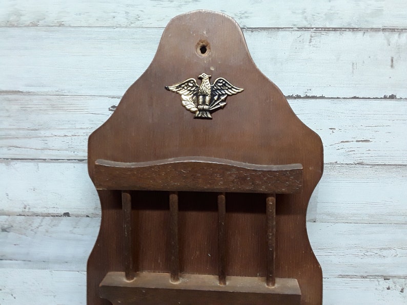 May include: Vintage wooden wall-mounted organizer with a decorative brass eagle emblem. Features a shelf and multiple slots for storage. The wood has a warm, brown tone, and the design is reminiscent of a classic, rustic style.