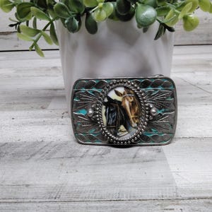Pode incluir: Fivela de cinto retangular prateada com um design turquesa e floral. O centro apresenta uma imagem oval de dois cavalos. A fivela tem uma estética vintage e ocidental.