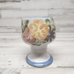 May include: A white ceramic goblet with a blue rim and a floral design painted in shades of yellow, orange, brown, and blue.