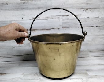 May include: A brass bucket with a black metal handle. The bucket is empty and has a worn, aged look.