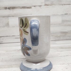 May include: A white ceramic mug with a blue handle and floral design. The mug has a pedestal base with a blue band around the bottom.