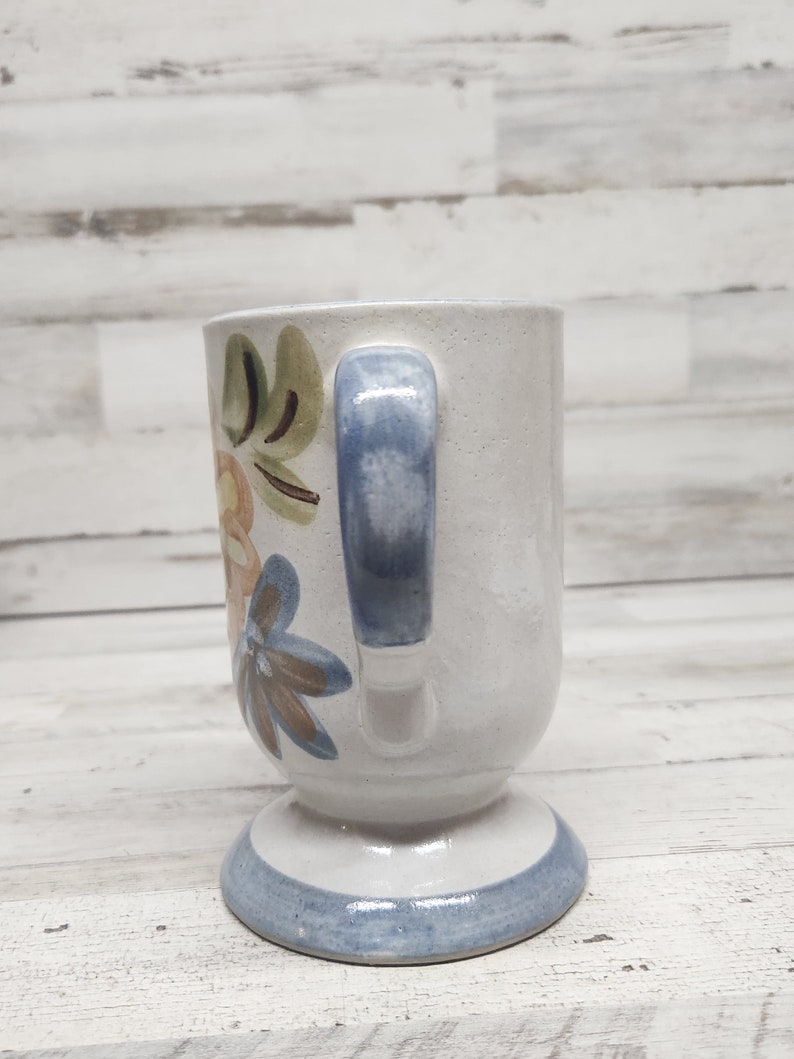 May include: A white ceramic mug with a blue handle and floral design. The mug has a pedestal base with a blue band around the bottom.