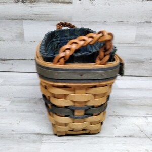May include: Woven basket with a braided leather handle. The basket has a two-tone design with natural and grey-green woven sections. The interior is lined with a blue and green patterned fabric. A decorative and functional storage item.