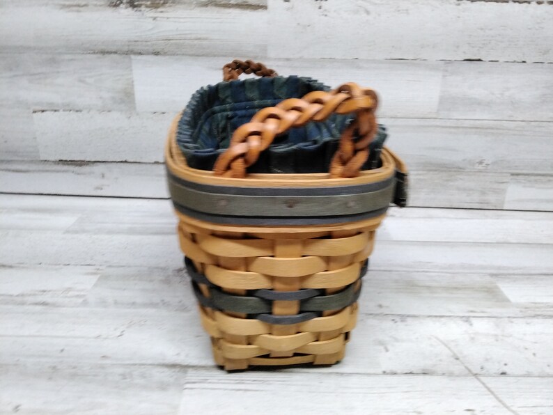 May include: Woven basket with a braided leather handle. The basket has a two-tone design with natural and grey-green woven sections. The interior is lined with a blue and green patterned fabric. A decorative and functional storage item.