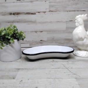 May include: A white ceramic rooster figurine, a white oval dish with a black rim, and a white ceramic pot with greenery. The objects are arranged on a light wood-grain surface.