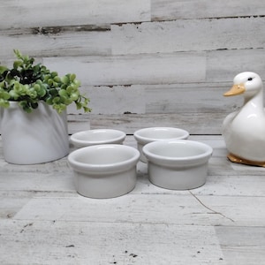 Op de afbeelding: Vier kleine, ronde, witte keramische ramekins staan op een licht houten oppervlak, naast een wit keramiek eendenbeeldje met een oranje snavel en poten. Een witte vaas met groen blad maakt de scène compleet.
