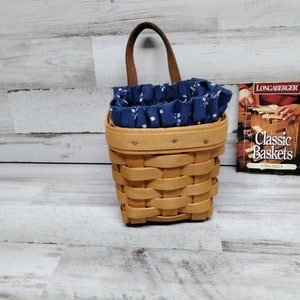 May include: A small, brown wicker basket with a blue and white fabric liner. The basket has a leather handle and is hanging on a wall. A small book titled "Classic Baskets" is visible in the background.